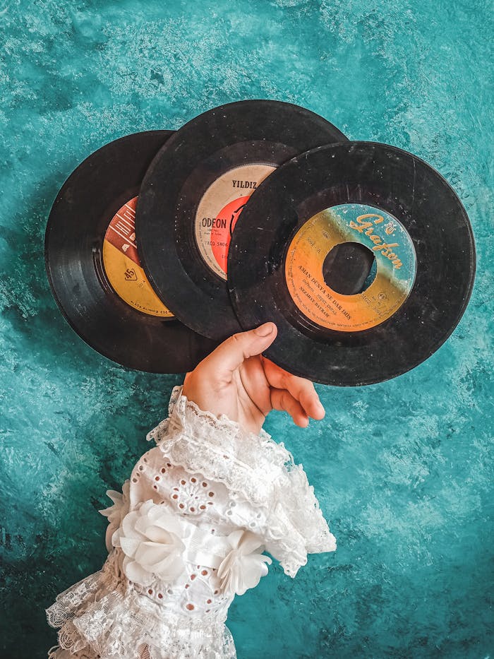 A hand in lace sleeve holds classic vinyl records against a teal backdrop.